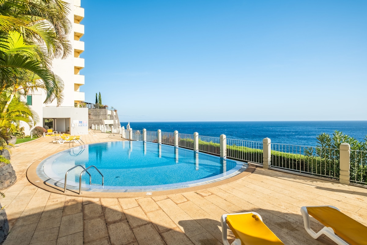 A curved, inviting swimming pool is set against the backdrop of the ocean. Sun loungers line the poolside, and palm trees add a touch of greenery. The clear blue sky reflects on the water, creating a serene atmosphere near the coastal edge.