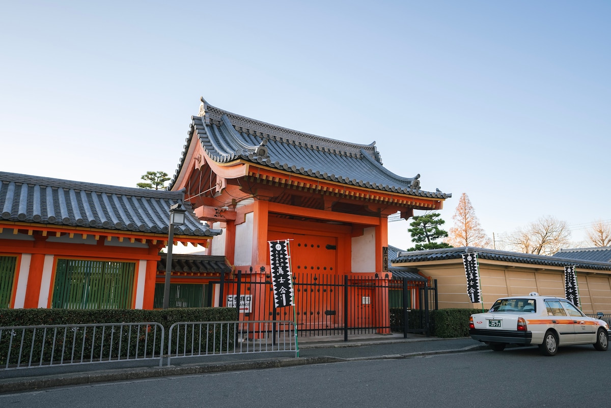 A traditional building with striking orange walls and intricate rooftops is visible, showcasing decorative features typical of Japanese architecture. Metal gates line the entrance, framed by lush greenery. A parked taxi is seen nearby, complementing the urban setting.