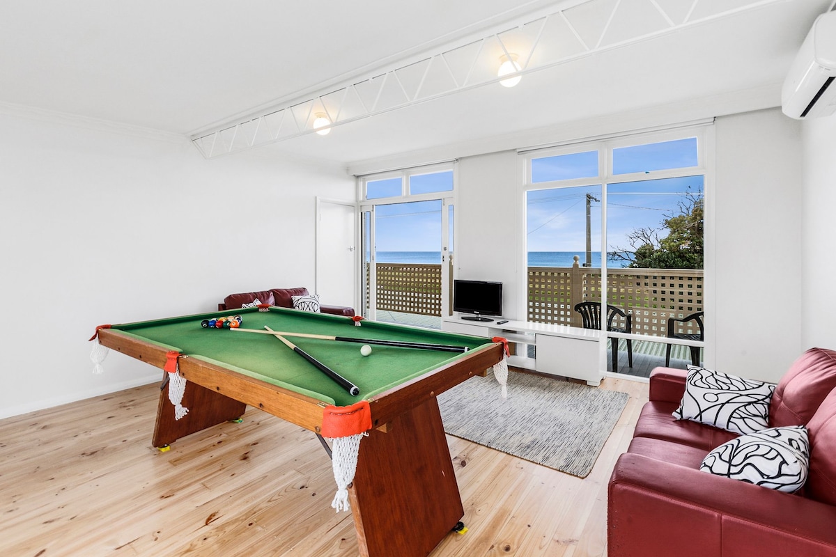 A spacious living area features a well-maintained pool table, complemented by two comfortable red sofas and a flat-screen TV. Large windows promote natural light and offer unobstructed views of the ocean, enhancing the inviting atmosphere of the space.