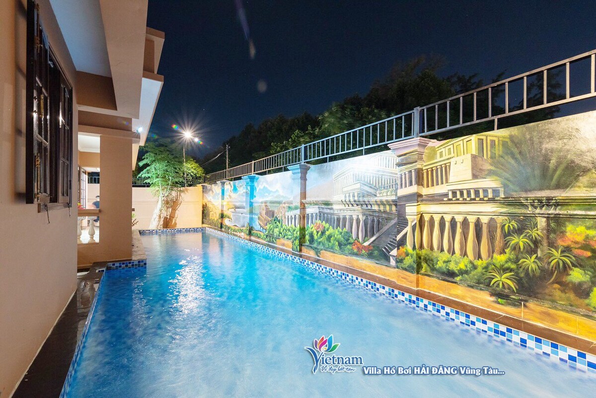 A swimming pool glistens under the night sky, surrounded by a mural depicting classical architecture. The calm water reflects the surrounding greenery, enhancing the serene atmosphere. Soft lighting illuminates the area, creating a tranquil setting for relaxation.