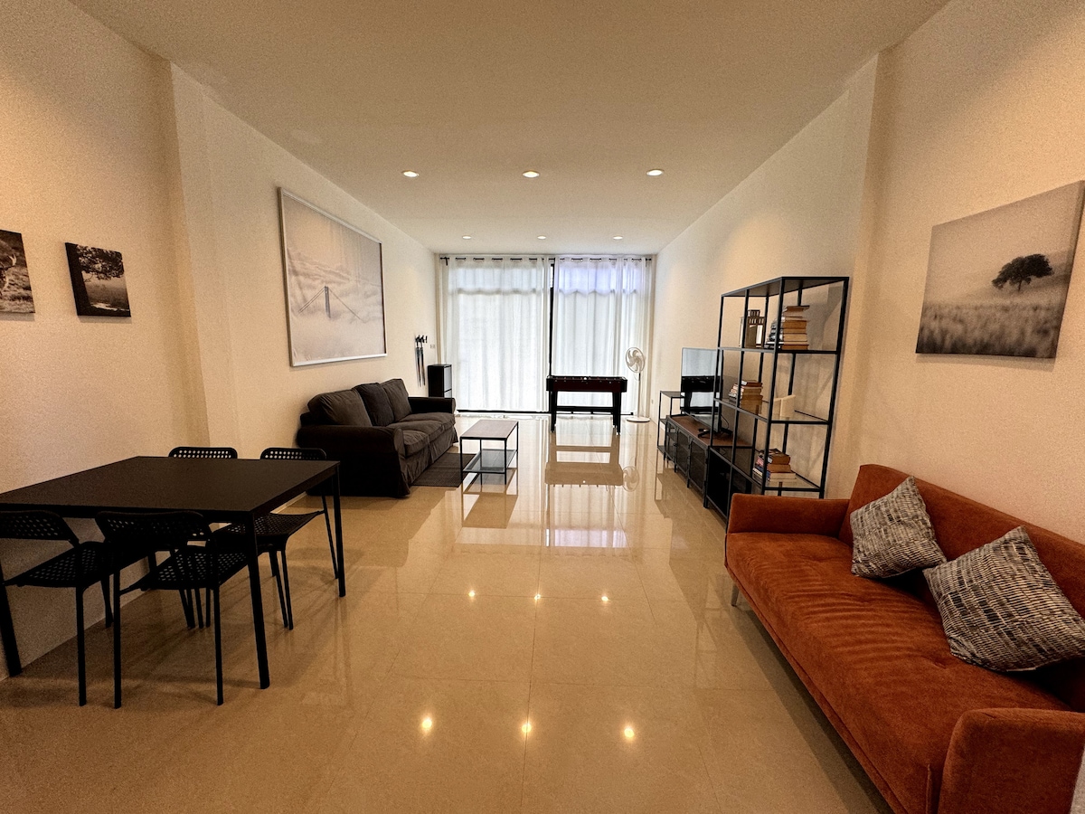 A spacious living room is presented, featuring a dark sofa and a vibrant orange couch complemented by decorative cushions. A dining table with black chairs occupies one corner, while a large window allows natural light to illuminate the polished floor. Minimalist decor is displayed on shelves.