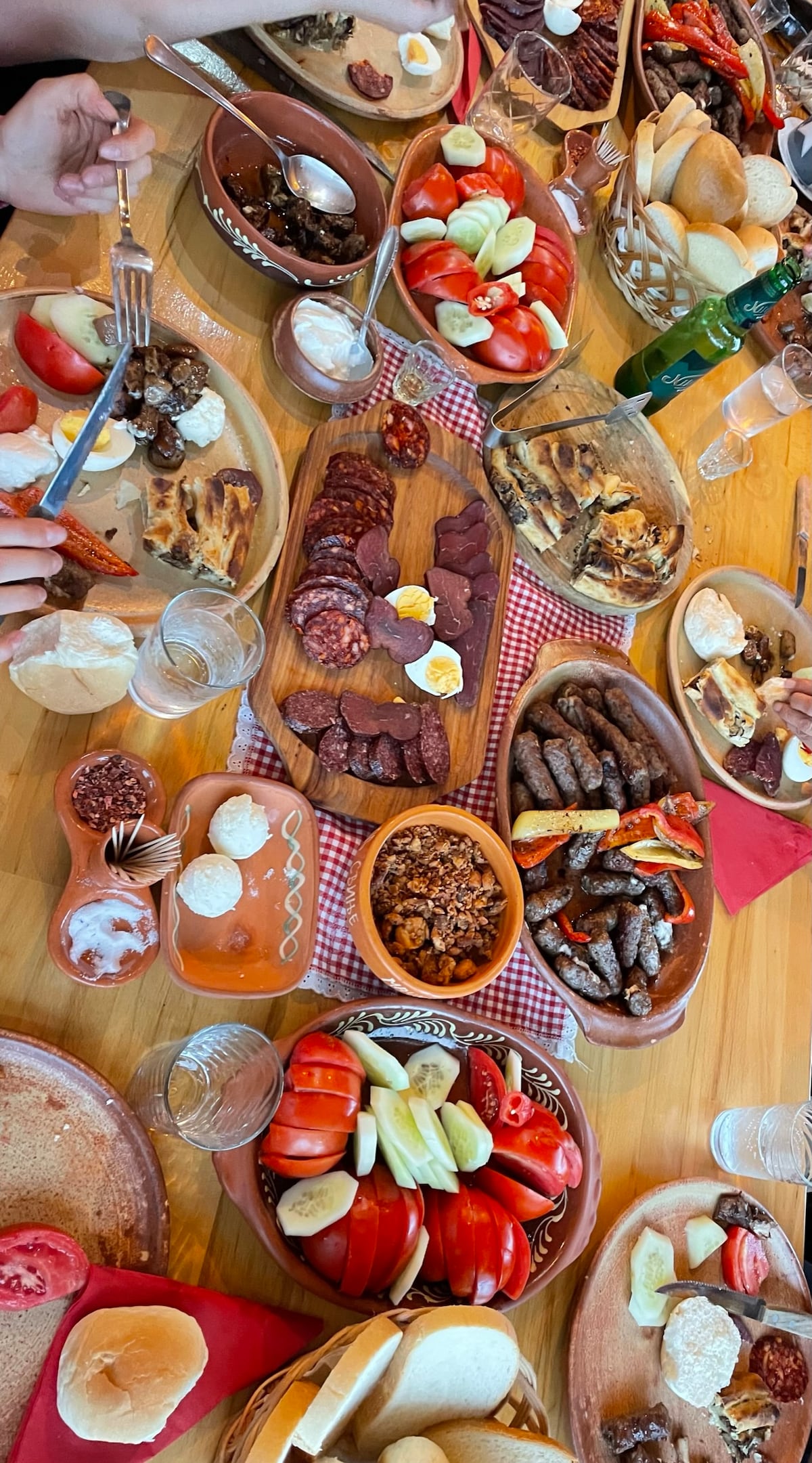 A wooden table is filled with various dishes, showcasing an array of colorful fruits, vegetables, meats, and cheeses. Plates of sliced tomatoes, cured meats, and grilled items are arranged alongside baskets of bread. Glasses of water add to the inviting dining setting.