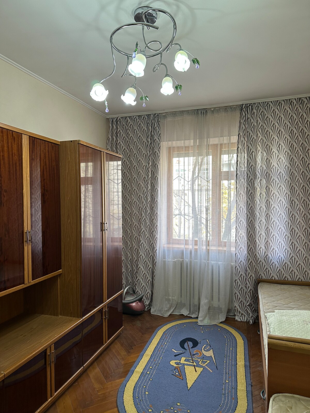 A well-lit bedroom is furnished with a combination of wooden cabinets and a spacious bed. A patterned rug is placed on the hardwood floor, and a large window features delicate curtains that allow natural light to filter into the room.