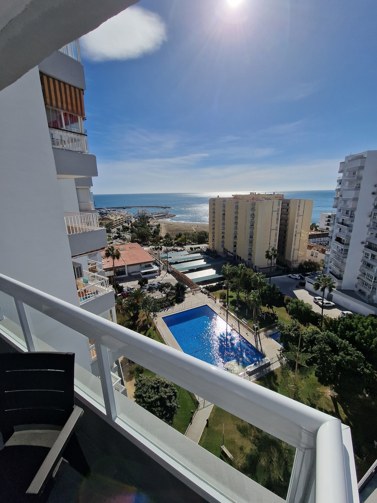 A sunlit balcony offers sweeping views of the coastline and the shimmering sea. Below, a swimming pool is surrounded by landscaped gardens. The scene includes nearby buildings and the vibrant atmosphere of the area, reflecting the outdoor leisure opportunities available.