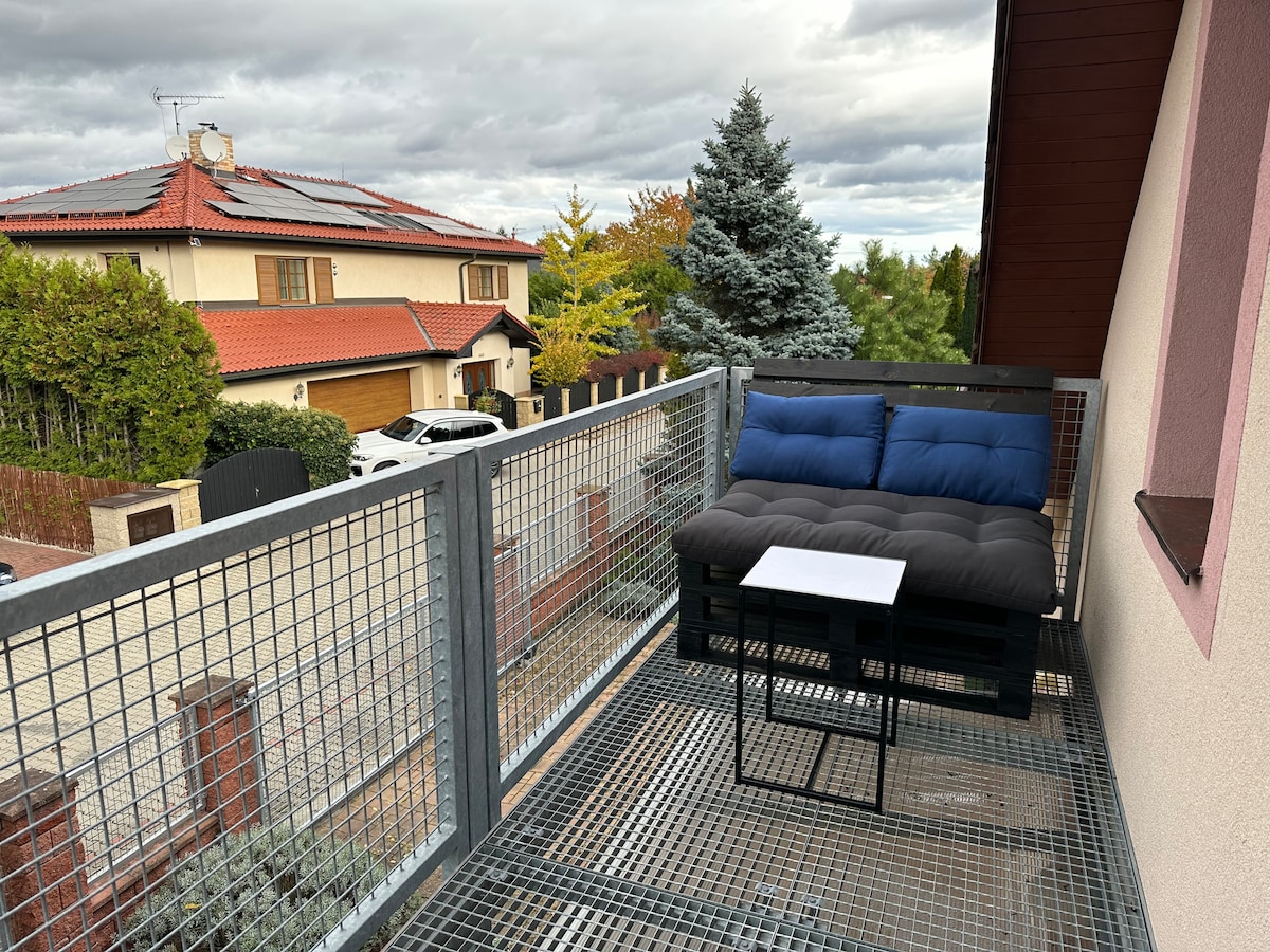 An outdoor terrace is present, featuring a blue cushioned seating area and a small table. Surrounding greenery and nearby homes are visible, with a cloudy sky providing a neutral backdrop.