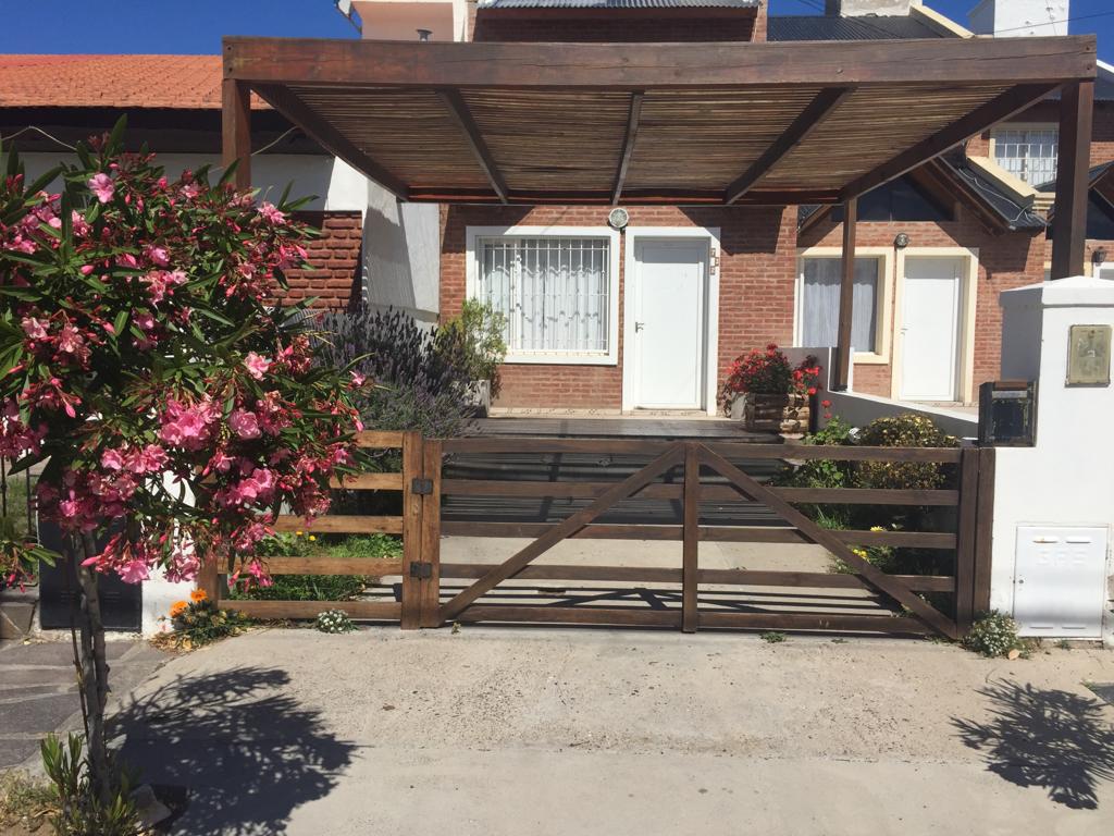 A modest duplex is framed by a wooden carport at the entrance. A wooden gate provides access, with a paved driveway leading to the front door. Flowers in front add touches of color, enhancing the inviting appearance of the home.