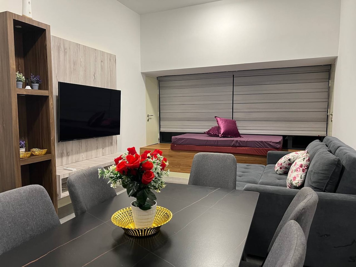 A modern living area features a plush gray sofa and a stylish dining table set for six. A wall-mounted television is positioned above a textured panel. A vibrant floral arrangement in a yellow bowl sits at the table’s center, while a daybed with a purple cushion is situated in the background.