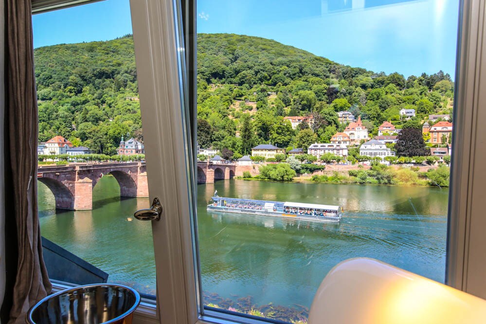 A scenic view is presented from the window, showcasing the river and a historic bridge. Lush green hills rise in the background, while a boat navigates the water's surface, enhancing the picturesque landscape.