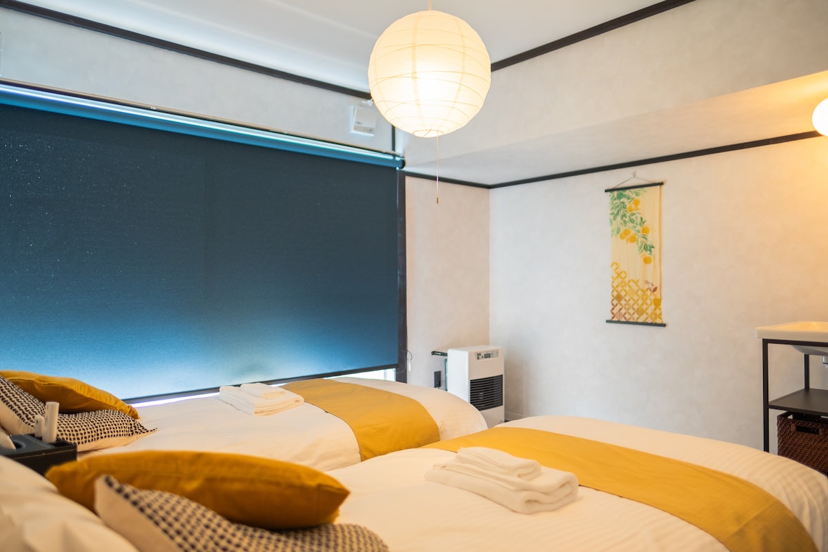 A cozy room is equipped with two spacious beds adorned with patterned throw pillows and warm blankets. A large window is covered with a dark roller shade, allowing for controlled light. Traditional wall art and a modern air-conditioning unit add to the inviting ambiance.