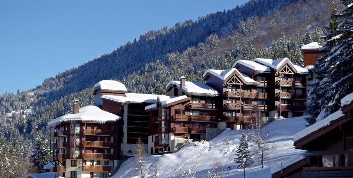 A cluster of modern buildings sits amidst a snowy landscape, with sloping roofs blanketed in white. Surrounding trees display a dusting of snow, contributing to the serene winter atmosphere. Mountain slopes rise in the background, creating a picturesque setting for outdoor activities.