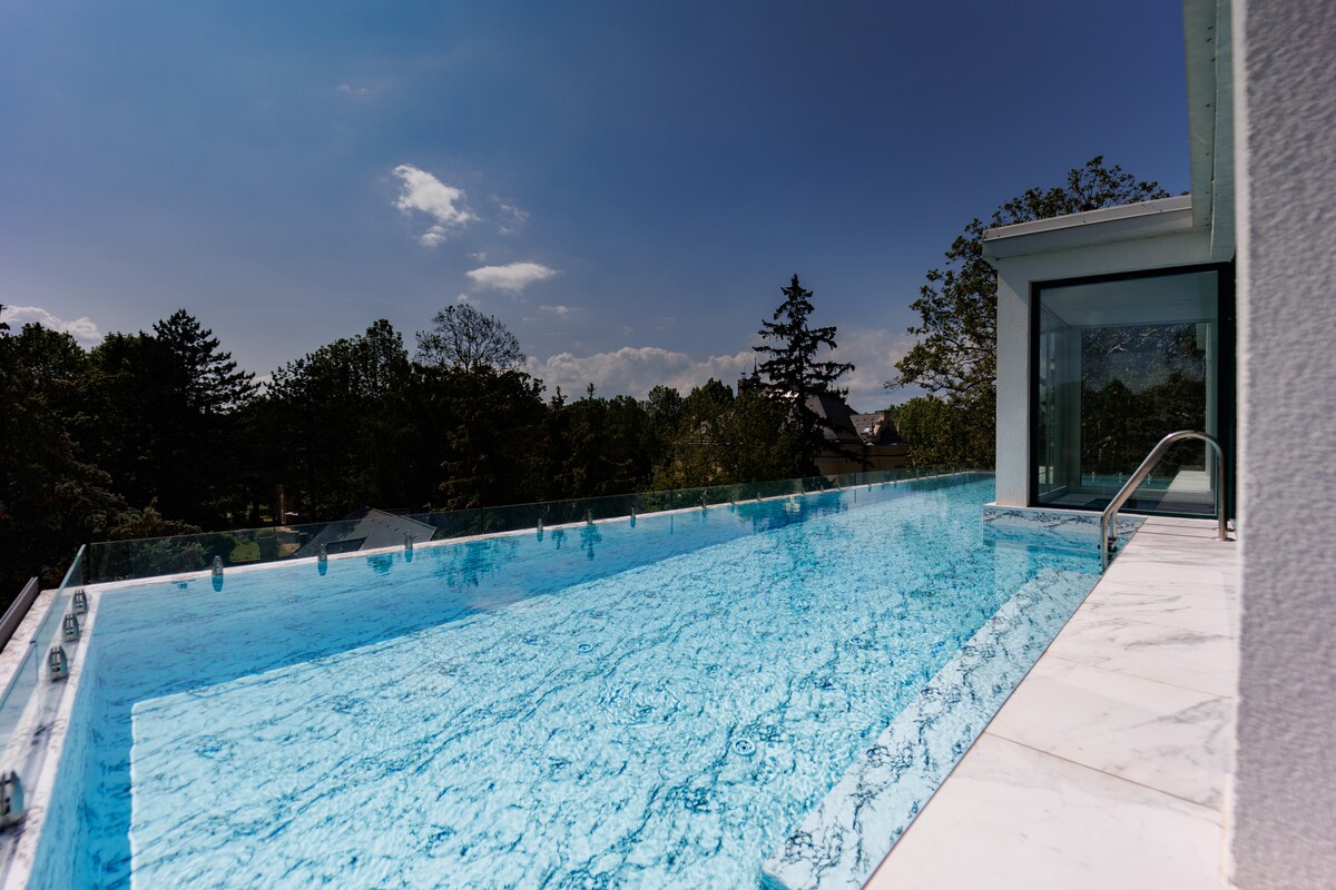 An infinity pool extends towards the horizon, surrounded by lush trees and a clear blue sky. The pool's glass edge reflects the light, providing an unobstructed view of the outdoors. Marble tiles line the deck, offering a sleek, modern aesthetic.