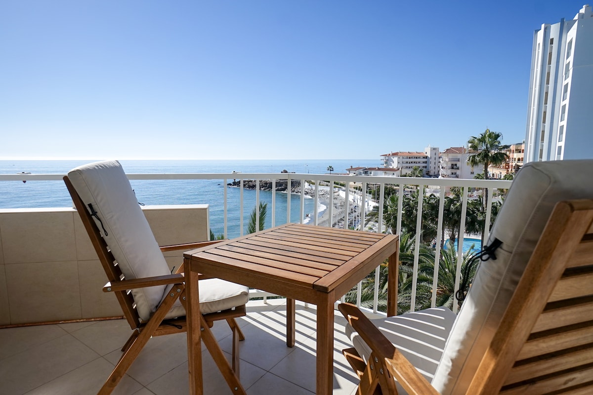 A balcony is depicted featuring a wooden table and two reclining chairs. The scene overlooks a shimmering sea, with the tranquil shoreline and palm trees visible below. A clear blue sky complements the coastal view, creating an inviting outdoor space.