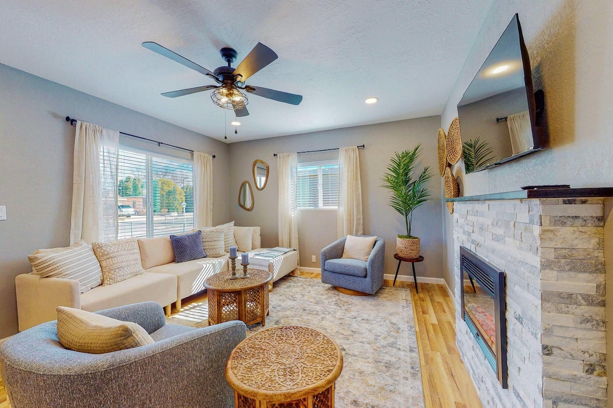 A welcoming living space features a comfortable sectional sofa and a cozy armchair, arranged around a coffee table. Natural light fills the room through large windows, and a decorative fireplace is framed by a stone accent wall. Plants and wall mirrors add to the ambiance.