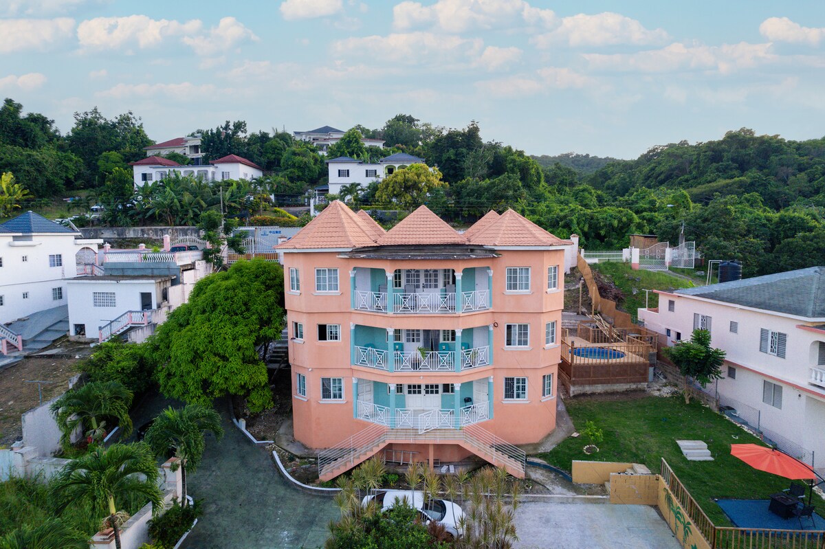 A three-story building features a vibrant peach exterior, complemented by a tiered staircase leading to spacious balconies. Lush greenery surrounds the property, while a large yard is visible alongside. An above-ground pool and gazebo are located in the backyard, enhancing outdoor leisure options.