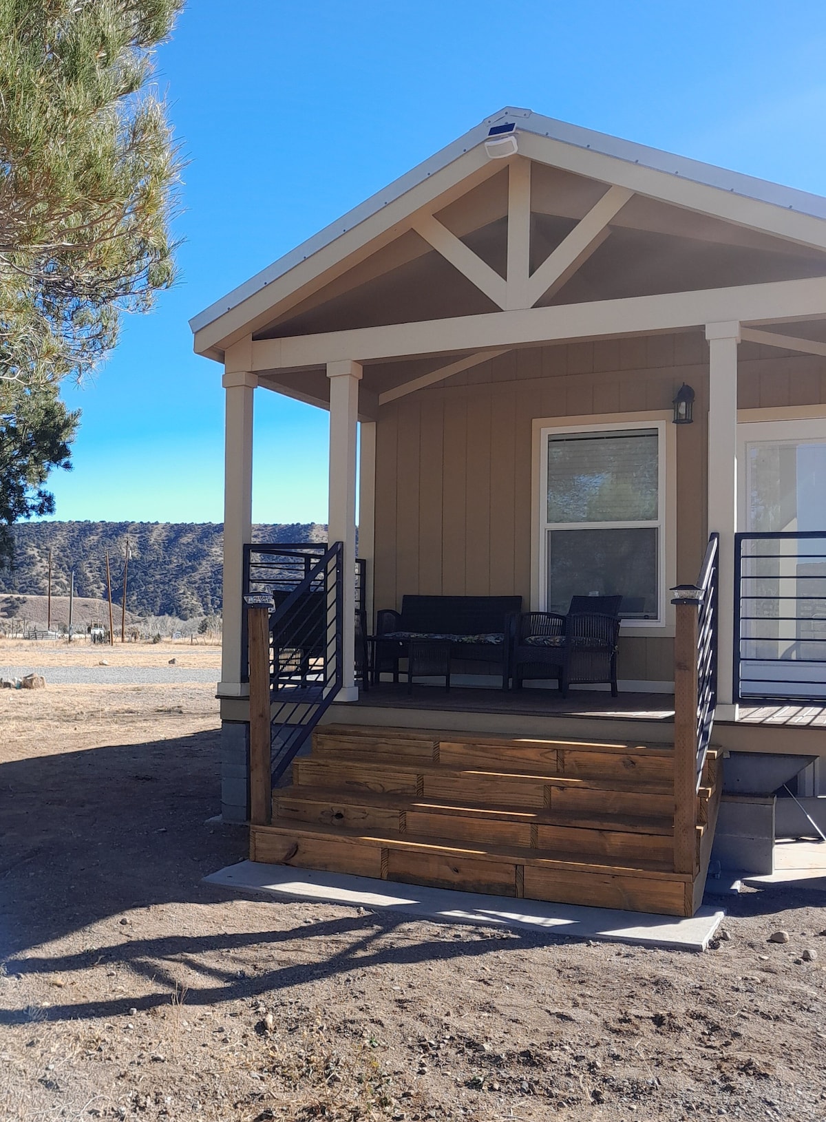 The exterior of a luxury tiny home is displayed, showcasing a covered porch with seating made of dark wicker. Sturdy steps lead up to the entrance, framed by a natural landscape. Sunlight illuminates the structure, enhancing its clean lines and inviting entrance.