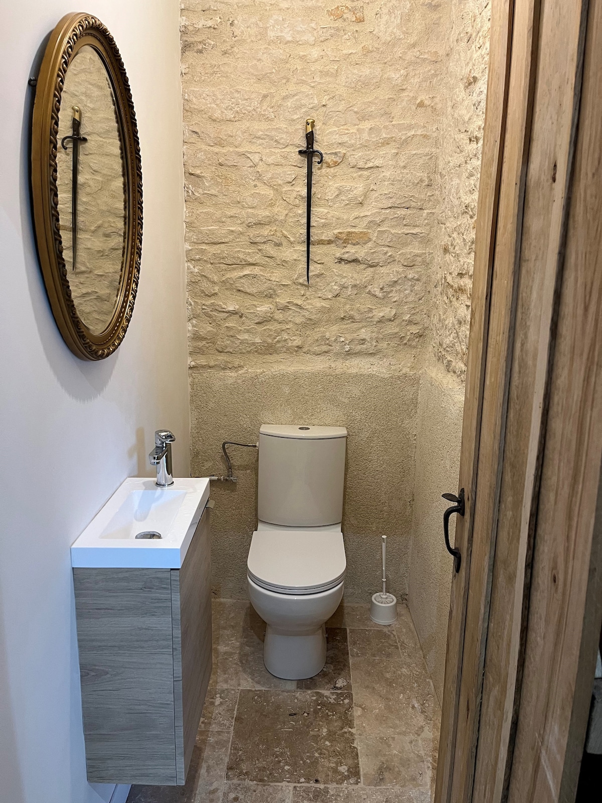 A compact bathroom features a modern toilet and a minimalist sink with storage beneath. A round mirror hangs above the sink, reflecting the stone wall. Natural stone tiles adorn the floor, contributing to the clean and functional design. A sword mounted on the wall adds a unique touch.