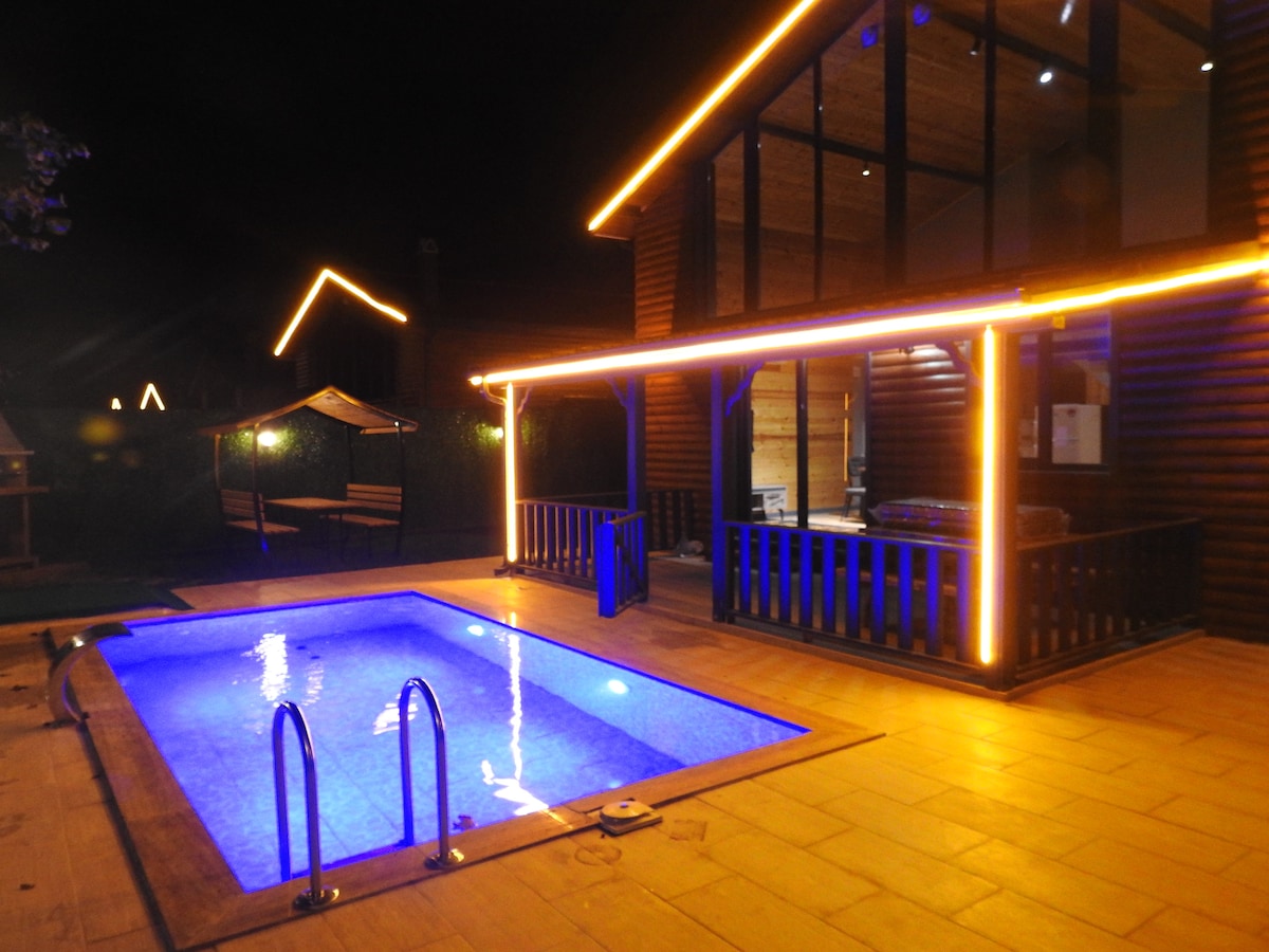An outdoor area is highlighted by a heated pool, illuminated with soft blue lighting. The wooden structure features warm lighting along the edges, providing an inviting ambiance. A covered sitting area can be seen nearby, adding to the comfort of the space.