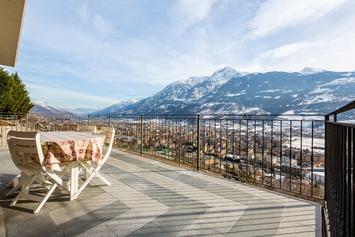 A spacious terrace with a dining set overlooks a panoramic view of Aosta and surrounding mountains. The snowy peaks contrast with the clear blue sky, creating a serene outdoor space for gatherings. The railing adds safety while enhancing the scenic experience.