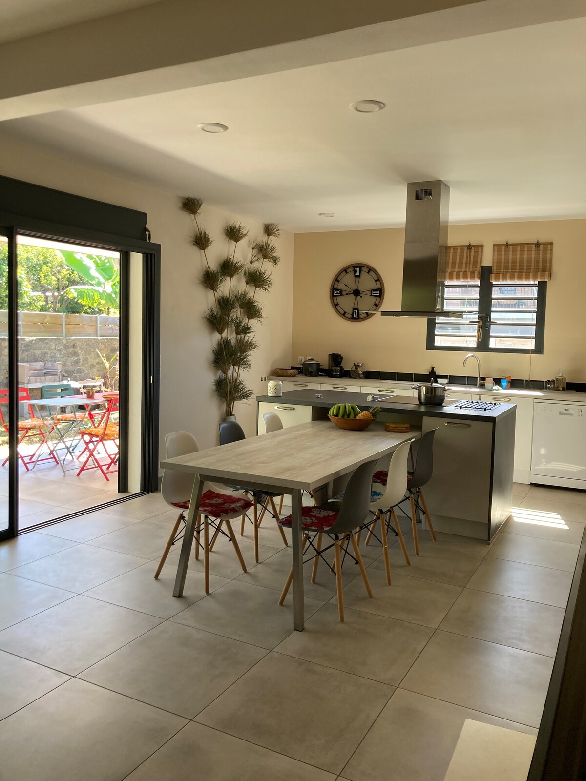 An open kitchen is showcased with a modern island and a dining table surrounded by colorful chairs. Natural light streams through large glass doors leading to an outdoor seating area adorned with red chairs. Decorative plants are placed along the wall for added interest.