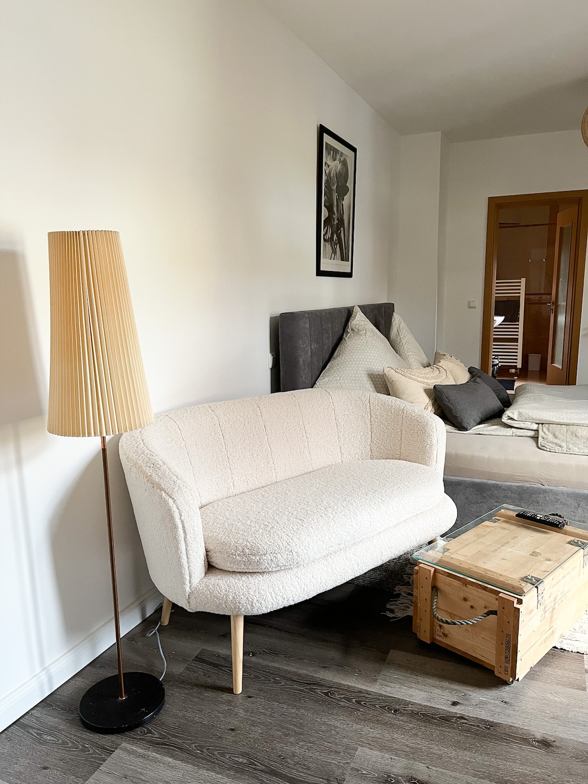A light-colored, plush sofa is positioned beside a stylish floor lamp featuring a ribbed shade. A wooden crate serves as a coffee table in front of a neatly made bed, which is decorated with cushions. The room is illuminated by natural light.