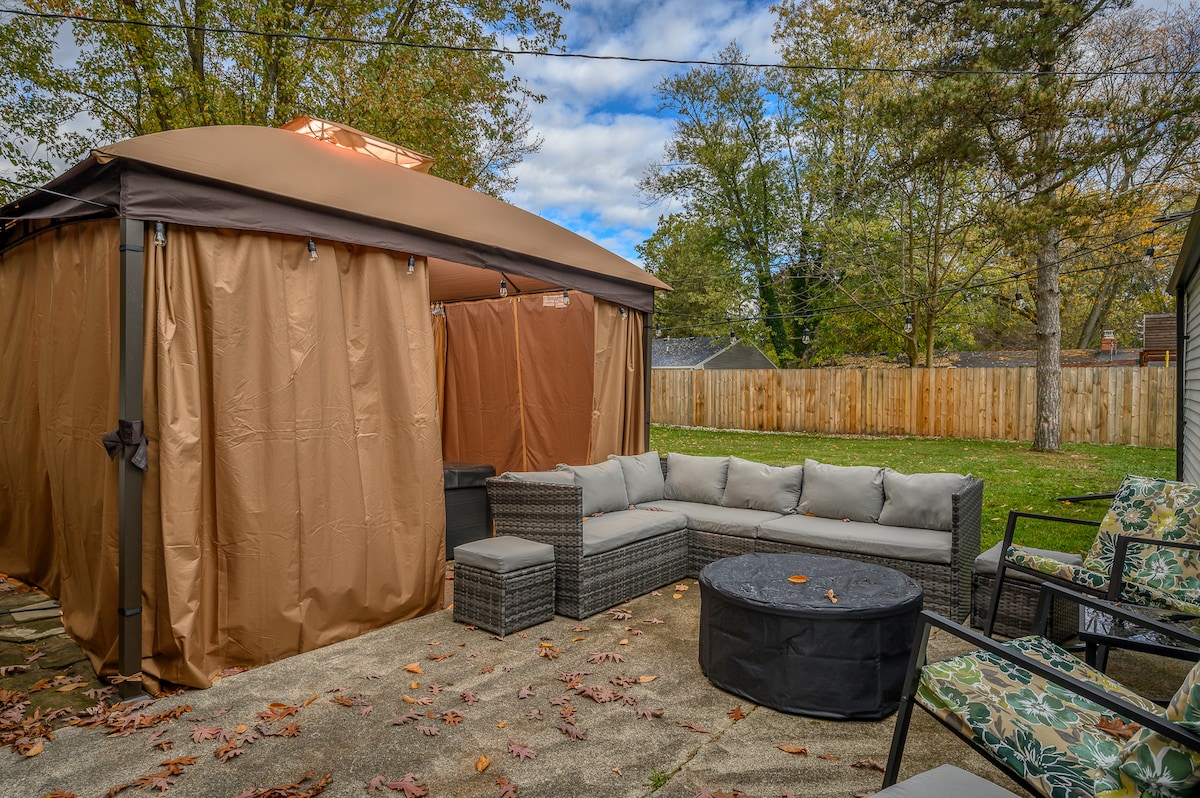 A gazebo encloses a hot tub, surrounded by comfortable seating, including a sectional couch and two armchairs. A round table sits in the center, creating an inviting outdoor space. The backyard features a fenced perimeter and is adorned with fallen leaves, indicating the season.