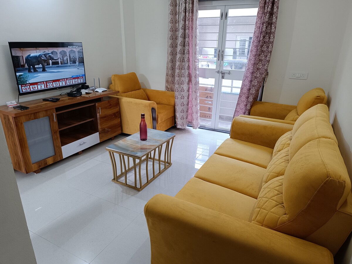 The living area features a spacious yellow sofa set paired with an accent chair, positioned around a glass coffee table. A television is mounted on the wall, and large windows allow natural light to fill the space, while decorative curtains add a touch of color.
