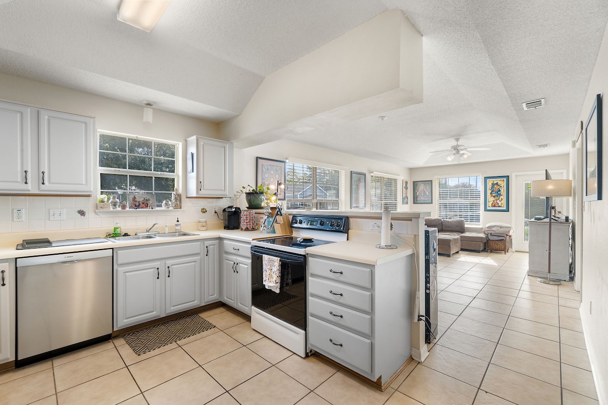 A bright kitchen features light grey cabinets and a spacious countertop. Stainless steel appliances, including a dishwasher and stove, are visible. Large windows allow natural light to fill the space, offering views of the living area, which includes comfortable seating and colorful artwork.