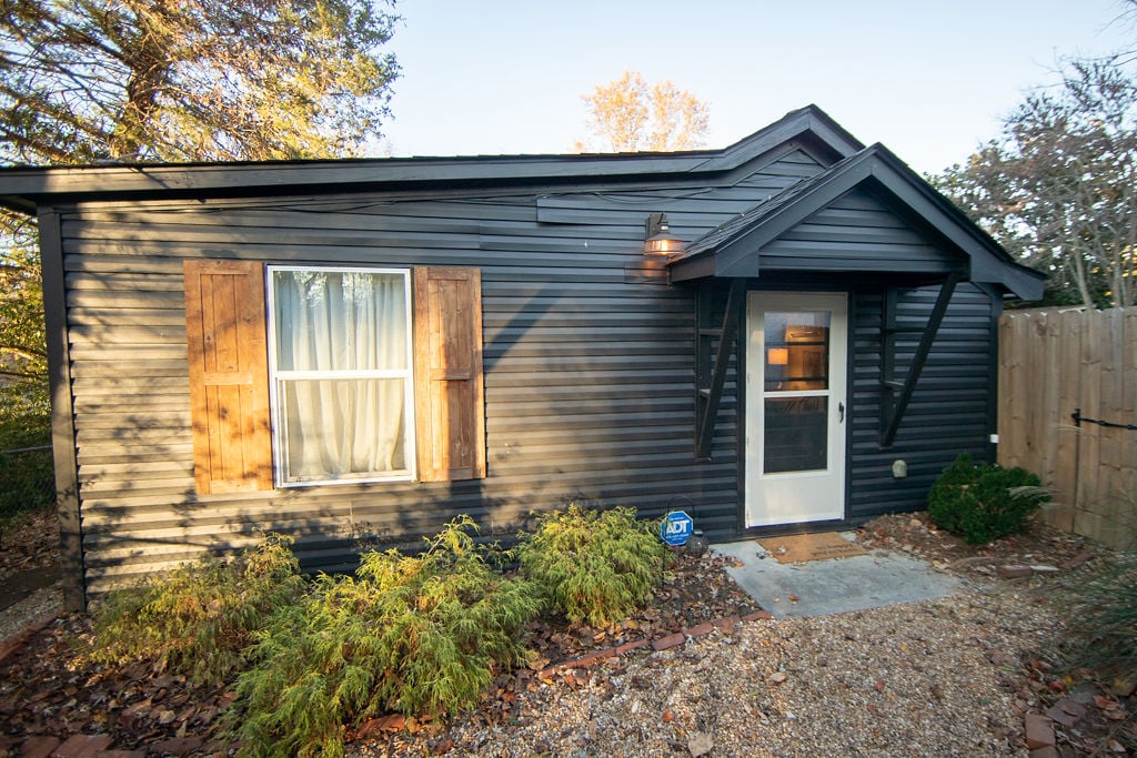 The exterior of the guesthouse features a modern black facade complemented by wooden shutters. A welcoming front porch with a simple mat leads to the main entrance. Surrounding greenery and gravel pathways enhance the inviting atmosphere.