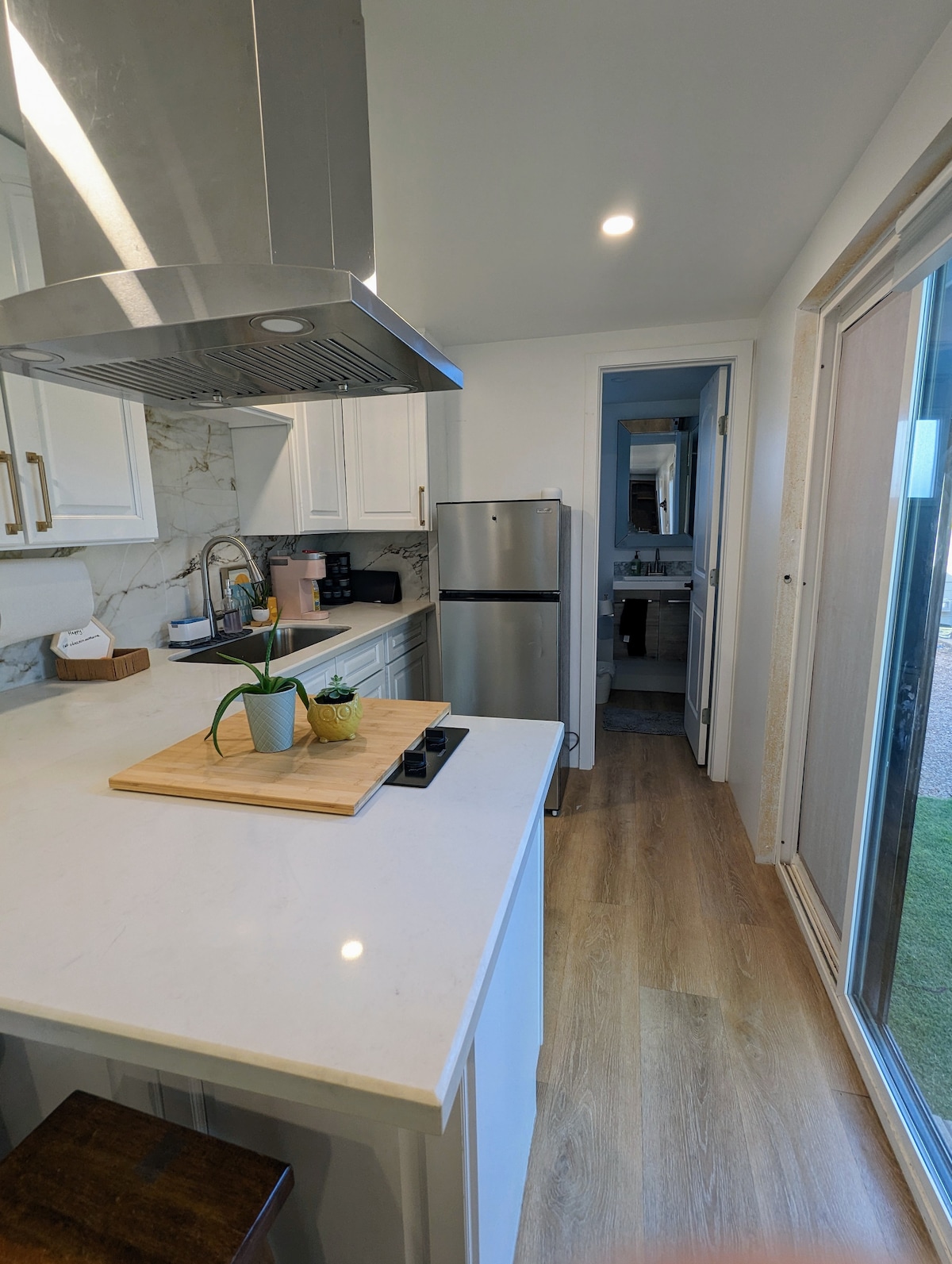A modern kitchenette is displayed with white cabinetry, a marble backsplash, and stainless steel appliances. A wooden cutting board with a plant adds a touch of greenery. A doorway leads to a full bathroom, with sliding glass doors providing access to the outdoor area.