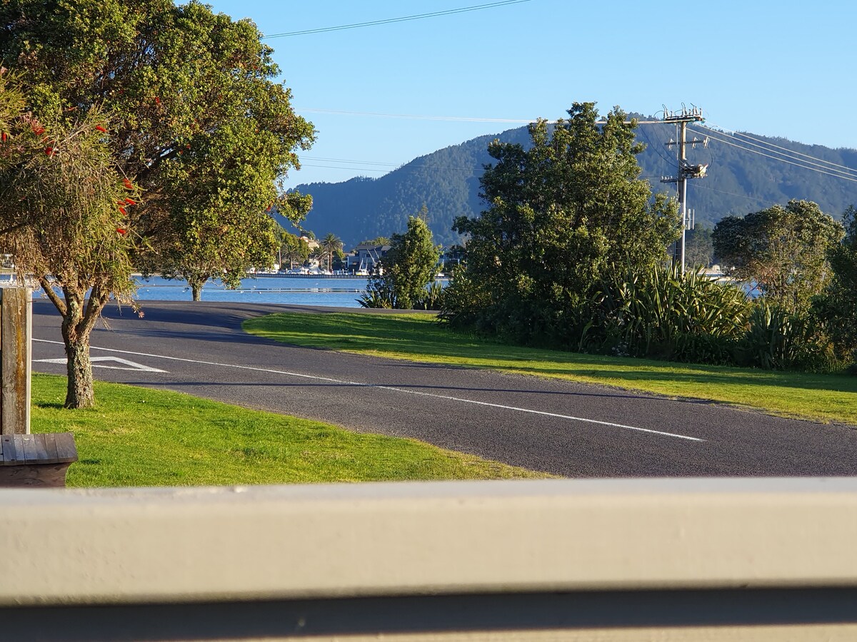 A scenic view captures a winding road bordered by lush greenery, leading towards a calm body of water. Distant hills stand majestically under a clear blue sky, enhancing the serene atmosphere of the surrounding landscape.