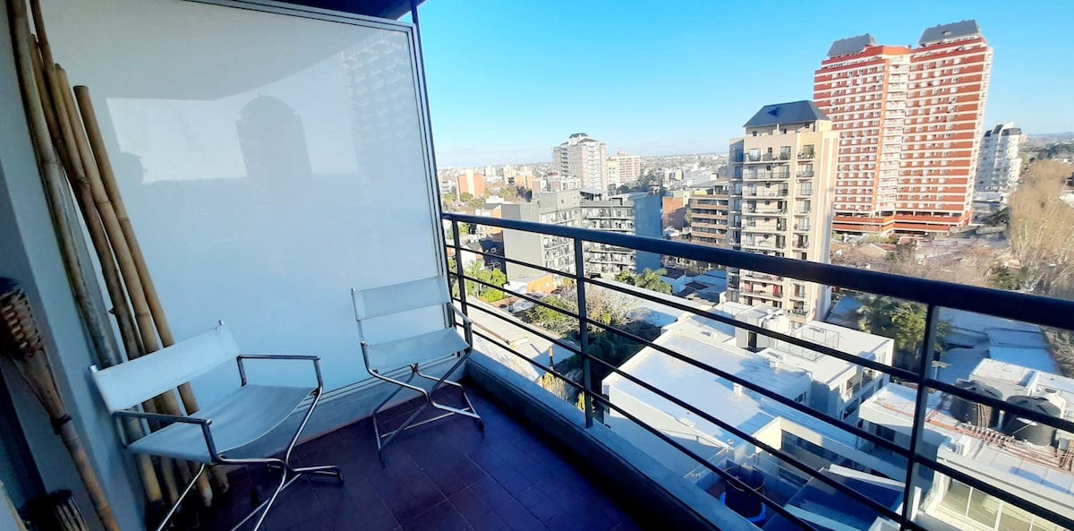 A balcony space is featured on the 10th floor, offering panoramic views of the surrounding cityscape. Two white chairs are positioned on the balcony, inviting relaxation, while a clear sky fills the backdrop.