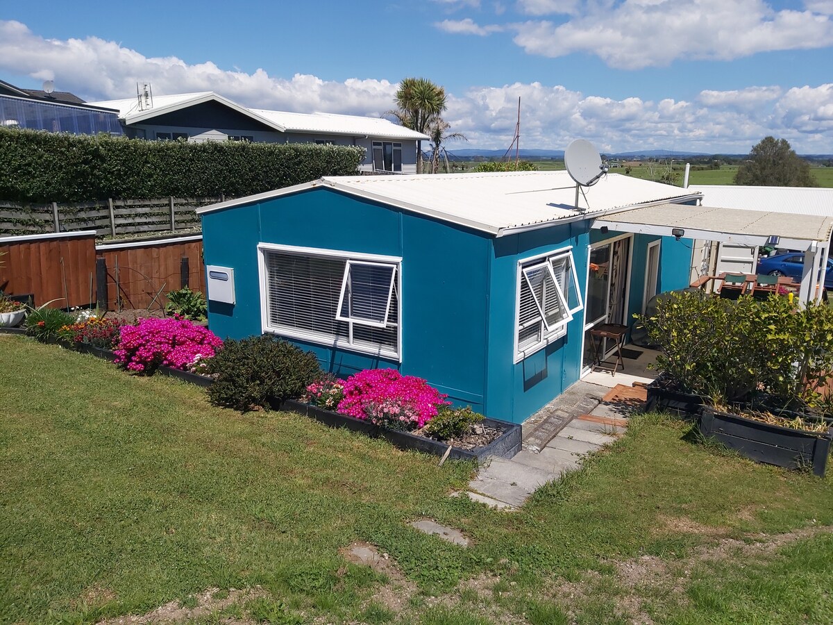A vibrant blue bach is framed by colorful flower beds, showcasing pink blooms and manicured greenery. The structure features multiple windows, allowing natural light to enter, and a covered outdoor area is seen at the side. Clear skies and distant fields provide a tranquil backdrop.