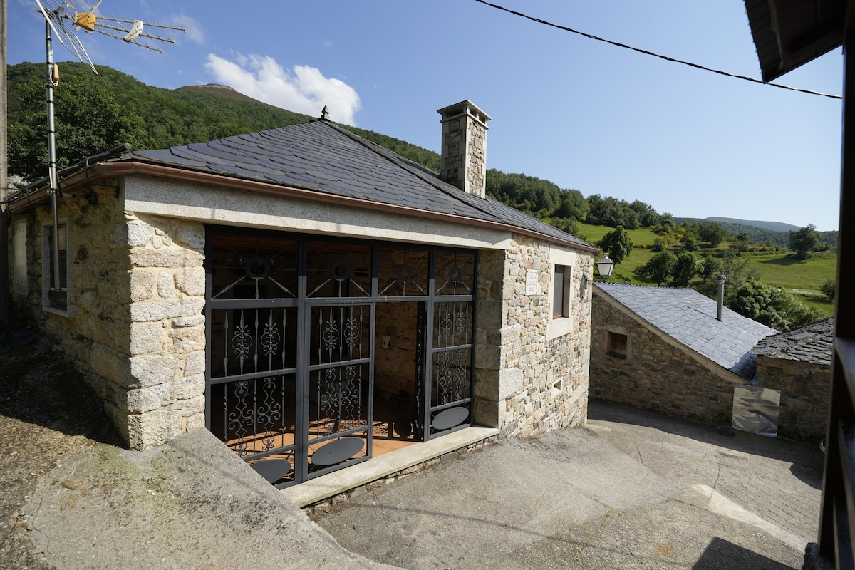 A rustic stone dwelling is displayed, featuring a sloped roof and a charming porch with wrought iron detailing. Surrounding green hills and trees create a serene backdrop. The driveway leads to additional nearby structures, enhancing the peaceful rural setting.