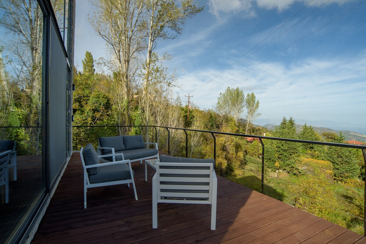 A private terrace is displayed with comfortable seating, featuring two armchairs and a side table. Lush greenery and distant mountains form a natural backdrop, while expansive glass doors reflect the outdoor scenery, inviting a blend of indoor and outdoor relaxation.