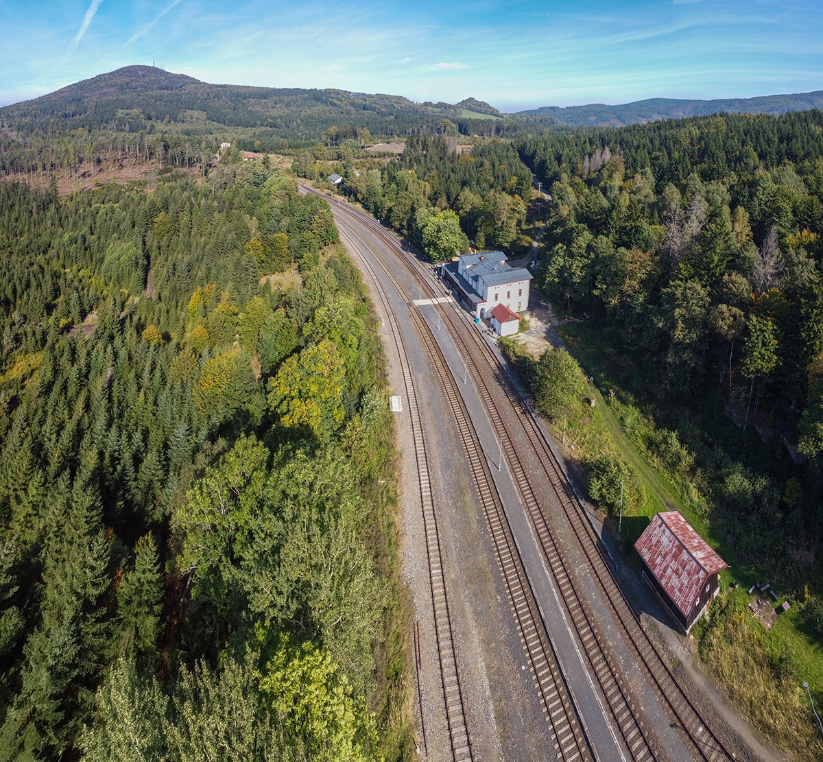 An aerial view captures the railway tracks winding through dense forests, with a building prominently positioned alongside. The surrounding greenery consists of tall trees and hills, creating a serene natural backdrop. A small red structure is visible near the tracks, indicating the rustic environment.