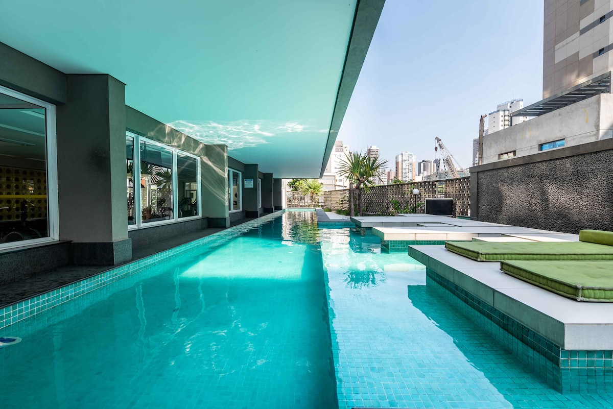 A serene swimming pool is surrounded by modern architecture, with crystal-clear water reflecting the sky. Comfortable lounge areas are situated beside the pool, complemented by greenery and open space, promoting relaxation in a tranquil urban environment.