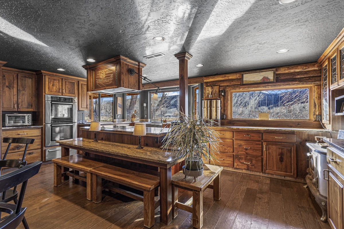 The spacious kitchen features warm wooden cabinetry and a large island with seating. Natural light is filtered through the windows, illuminating the wood accents. A minimalist dining area is present, enhancing the open and functional layout of the space, ideal for shared meals.