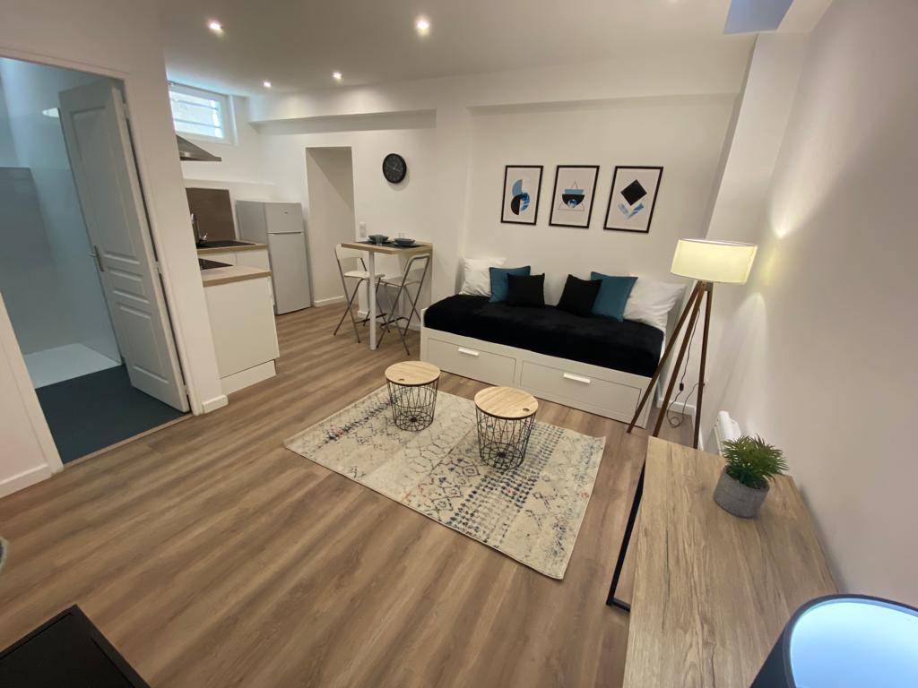 A well-arranged studio space features a cozy sleeping area with a dark sofa bed. A small dining area is visible, with a bar table and two high chairs. A modern kitchenette can be seen in the background, complemented by an inviting area rug and stylish light fixtures.