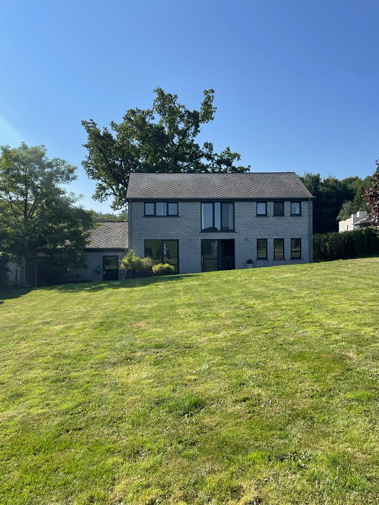 A light-colored house stands in a spacious, green lawn under clear blue skies. Large windows offer views of the surrounding area. A tree provides shade nearby, enhancing the serene outdoor environment.