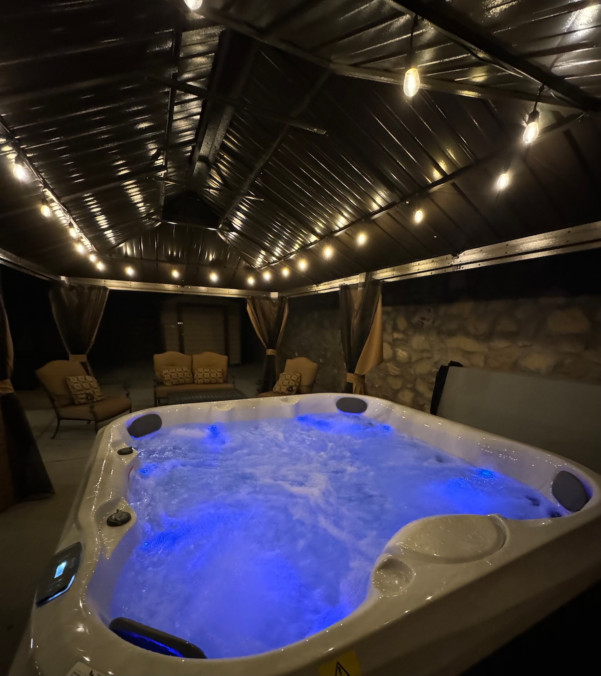 The outdoor jacuzzi is surrounded by a covered area with ambient lighting. The walls feature stone accents, and two cozy seating options are positioned nearby. Privacy curtains and a roof enhance the secluded atmosphere, while soft blue lighting illuminates the water, providing a calming ambiance.