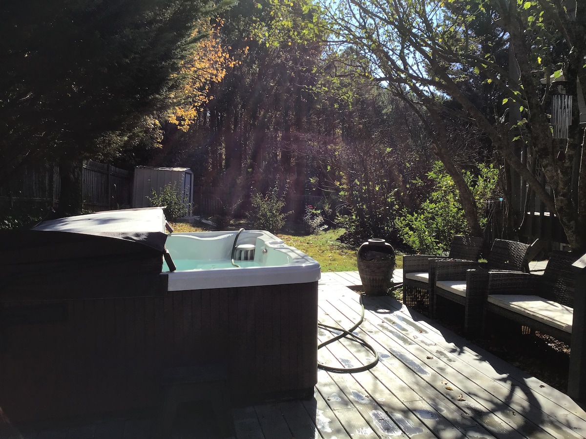 A private backyard is visible, featuring a hot tub on a wooden deck surrounded by lush greenery and sunlight. Comfortable seating arrangements are positioned nearby, inviting relaxation among the serene outdoor setting. Natural elements and trees frame the space, enhancing the tranquil atmosphere.