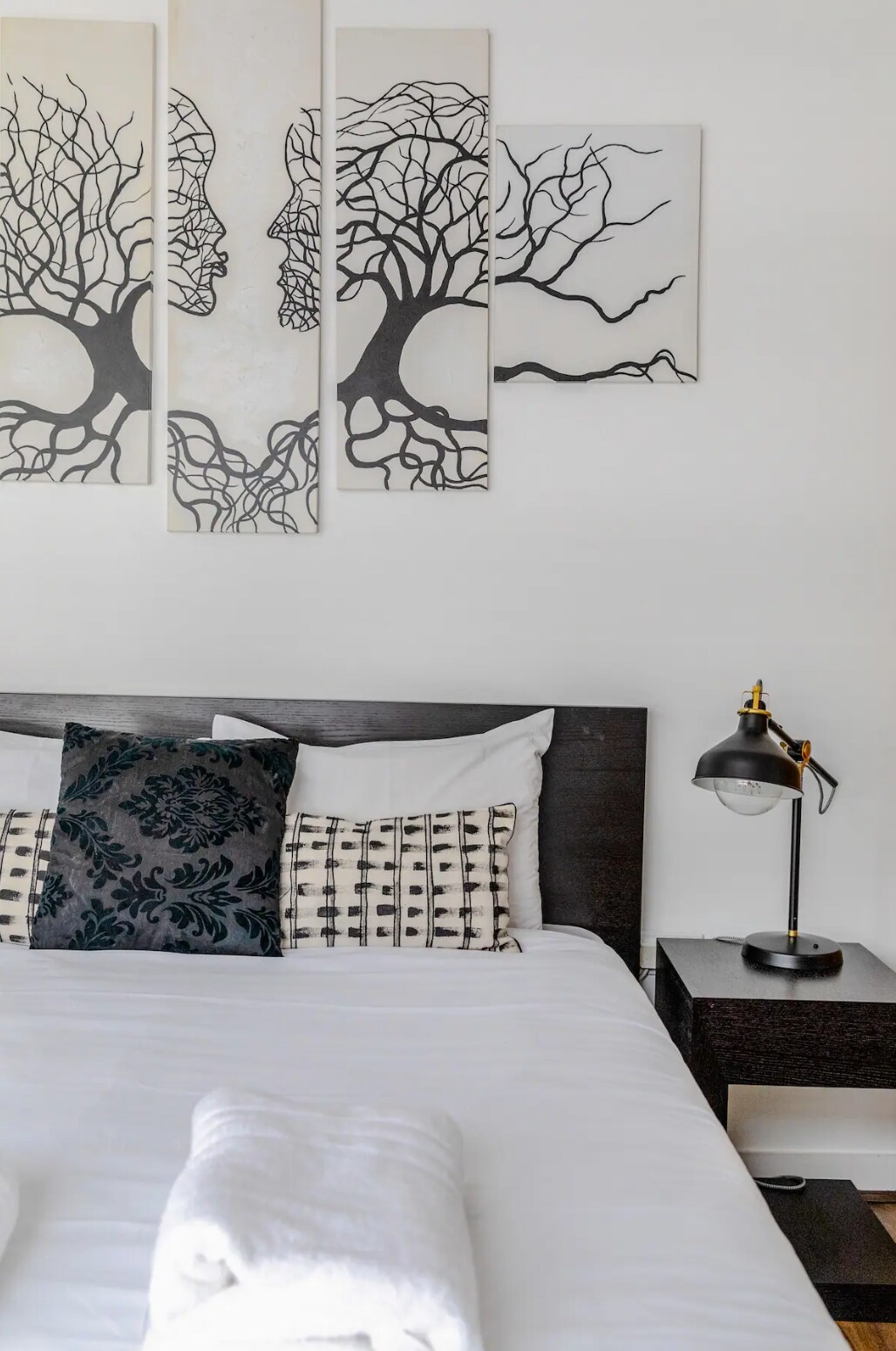 A spacious bedroom features a double bed with hotel-quality linens, complemented by patterned accent pillows. Above the bed, three art pieces depict intertwined faces and trees. A stylish bedside lamp and a dark nightstand complete the inviting setup.