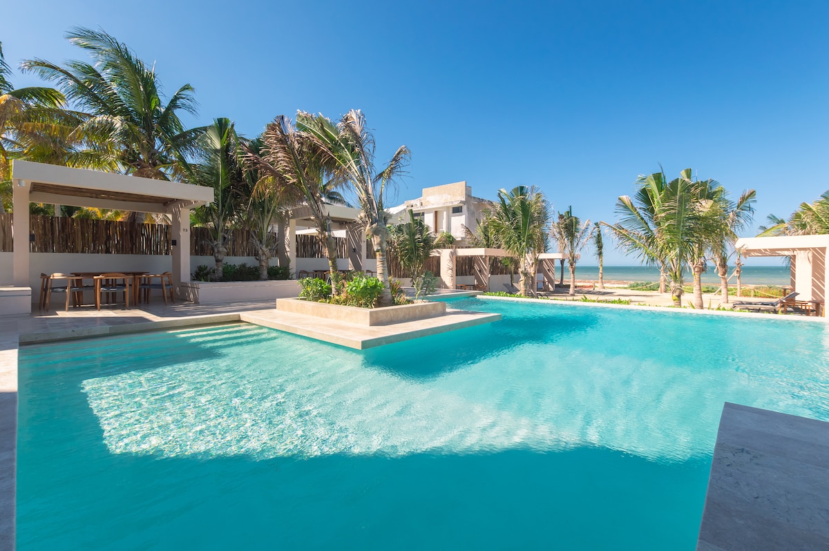 A large outdoor pool is set against a clear blue sky, surrounded by lush palm trees. Relaxation areas with shaded seating are visible along the pool's edge, providing a tranquil environment. The ocean horizon is faintly visible in the background.