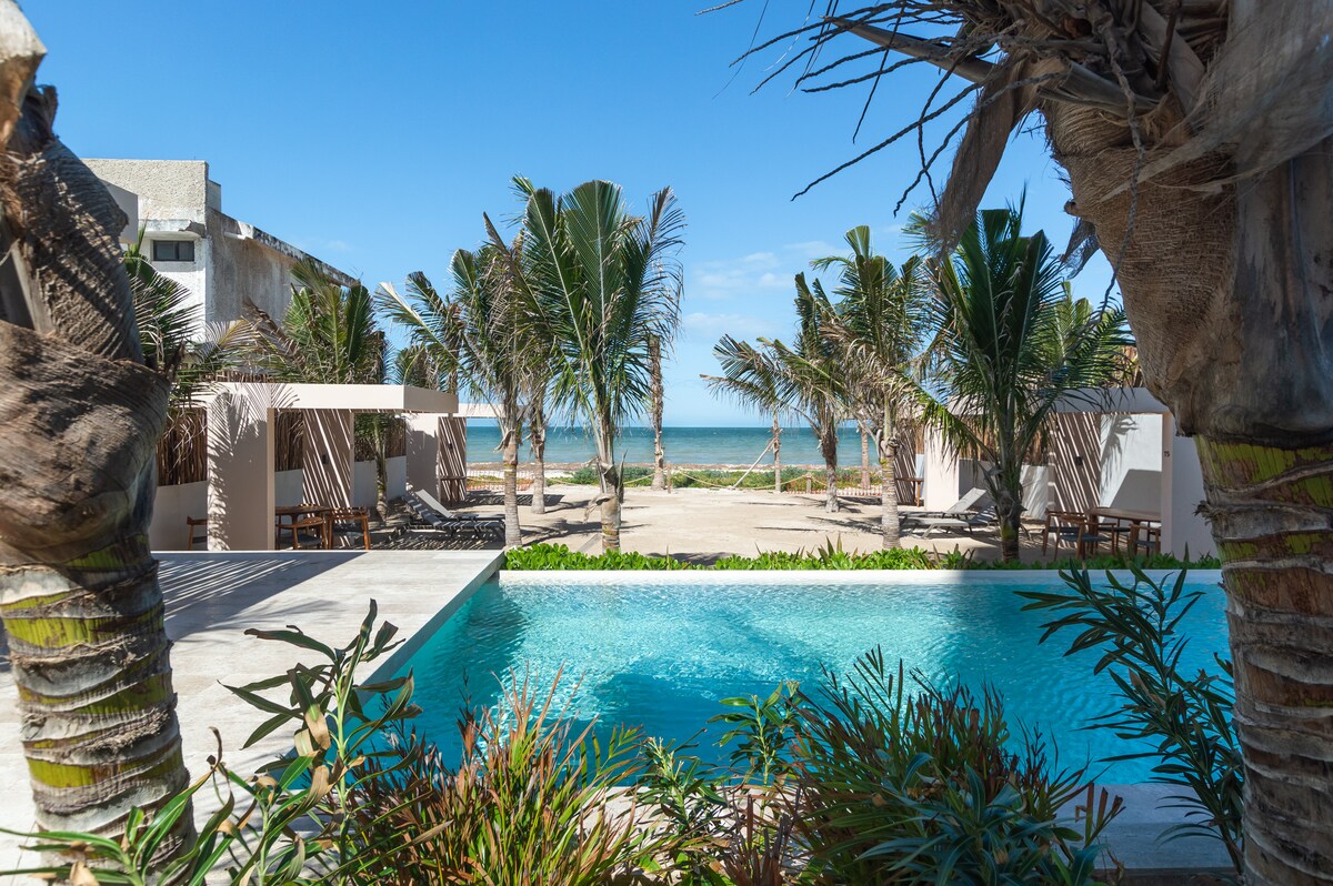 A tranquil view is presented from a patio, showcasing a swimming pool with clear blue water surrounded by lush greenery. Palm trees frame the scene, leading to a sandy beach and the calm ocean in the background under a clear blue sky.