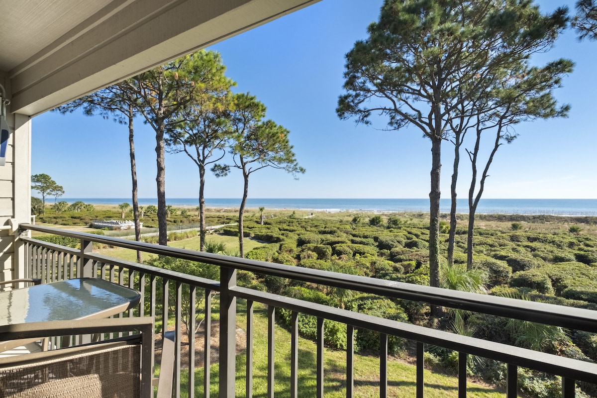 A private balcony offers elevated views of lush greenery and the ocean beyond. Tall pine trees frame the scene, creating a natural border against the clear blue sky. A table and chairs are positioned on the balcony, inviting outdoor relaxation.