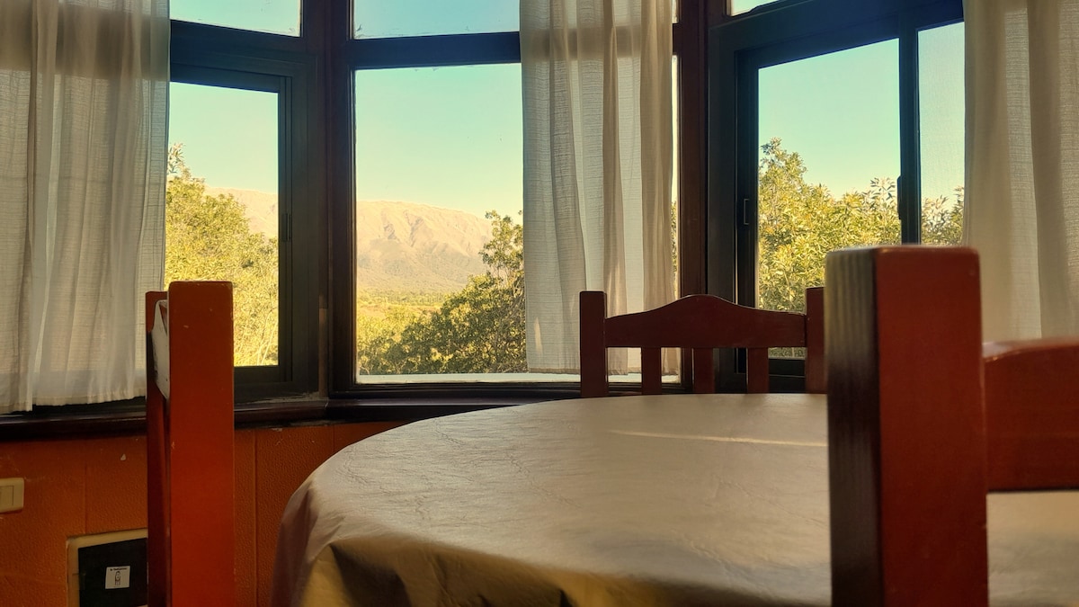 A dining area is presented with a round table and wooden chairs. Sunlight streams through large windows adorned with sheer curtains, creating a bright ambience. The view reveals natural landscape features, including rolling hills in the background.
