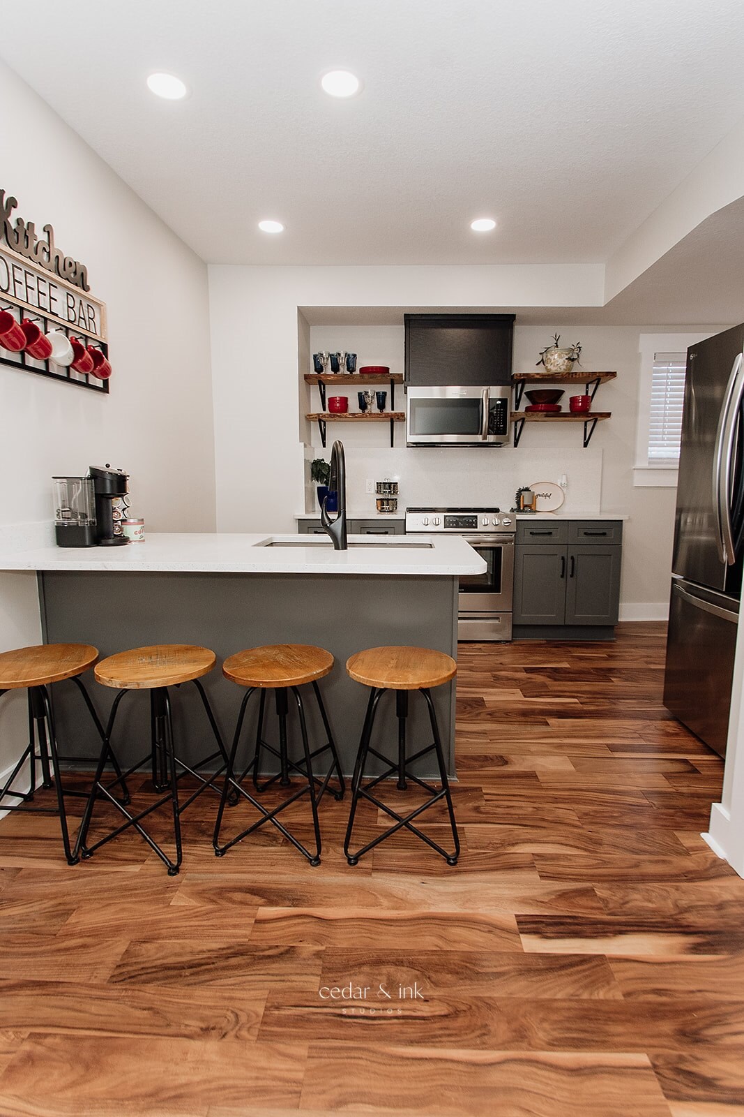 A modern kitchen features a sleek countertop with four high stools. Open shelving displays colorful dishware, while stainless steel appliances are neatly integrated. Wood flooring enhances the inviting atmosphere, and soft lighting highlights the workspace.