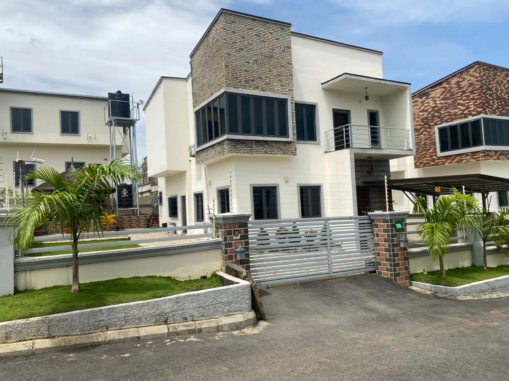 A modern duplex is presented with a striking blend of white and stone accents, featuring large windows that allow ample natural light. A manicured lawn with palm trees enhances the welcoming entrance, while a secure gate provides accessibility to the property.