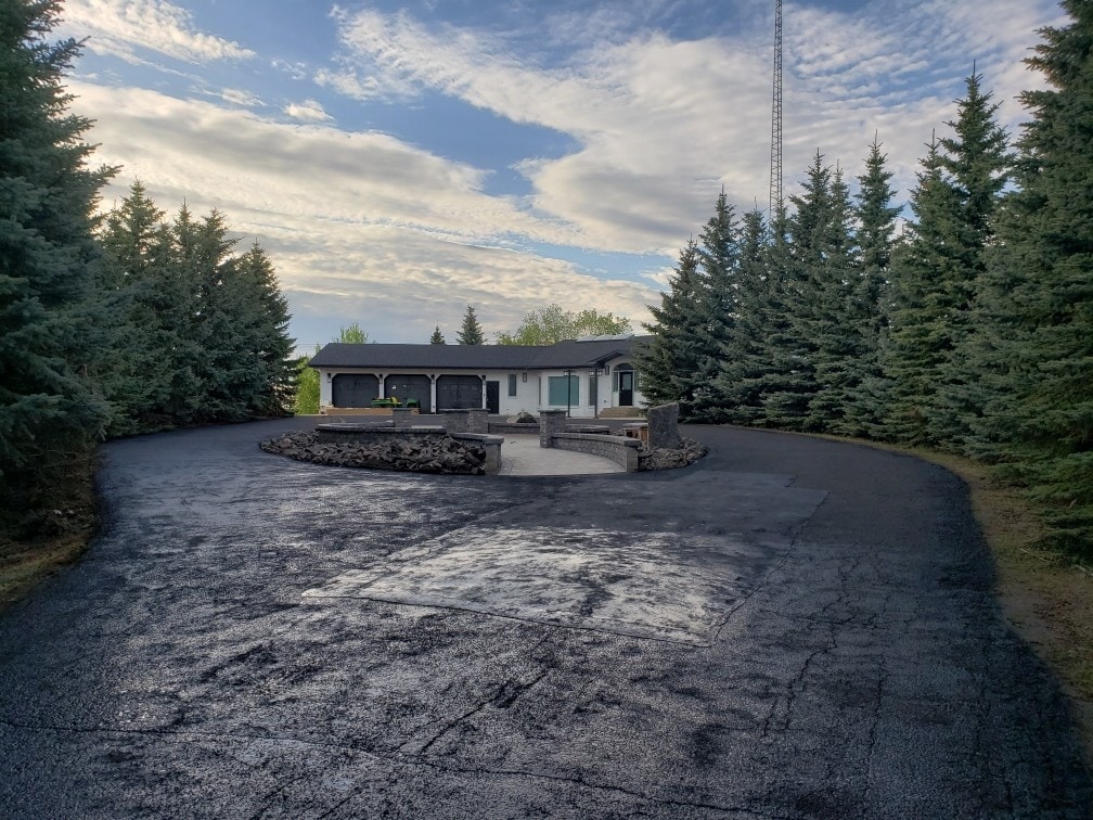 A spacious driveway winds through tall evergreen trees, leading to a modern home with a light exterior. Stone features outline a circular patio area, providing a welcoming entrance to the property. A clear sky with scattered clouds creates a serene backdrop.