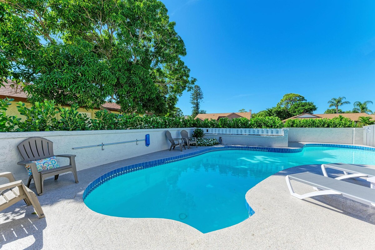 The image displays a private heated pool surrounded by lush greenery. Comfortable loungers are placed poolside, providing a serene space for relaxation. The clear blue water is inviting, reflected under bright sunlight, creating a refreshing outdoor atmosphere.